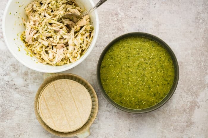 A bowl of shredded chicken mixed with green sauce, a bowl of green salsa, and a stack of corn tortillas on a light textured surface.