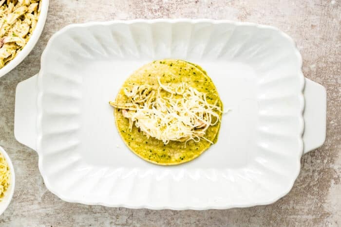 A corn tortilla filled with shredded chicken and cheese sits in the center of a white rectangular baking dish. The background is a light, textured surface with part of a bowl visible on the left.