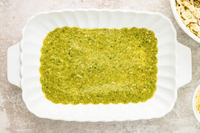 A white scalloped baking dish with a layer of green sauce spread evenly on the bottom, set on a light textured surface. A bowl with shredded ingredients is partially visible in the top right corner.