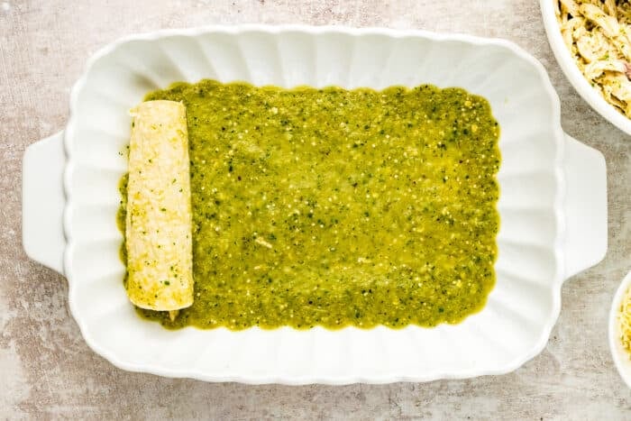 A white baking dish with green enchilada sauce spread on the bottom and one rolled enchilada placed on the left side; bowls with filling ingredients are partially visible nearby.