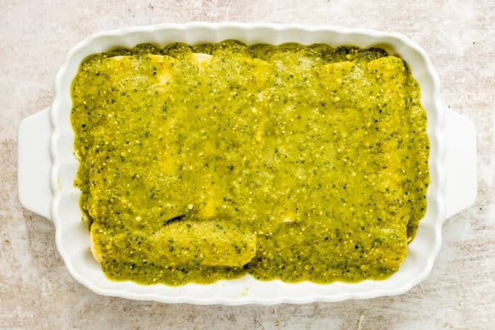 A white baking dish filled with rolled tortillas covered in a green salsa or sauce, likely enchiladas, on a light-colored surface.