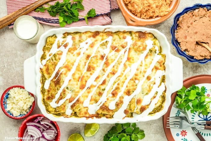A casserole dish of enchiladas topped with melted cheese and drizzled with crema, surrounded by refried beans, Mexican rice, sliced red onions, lime wedges, cilantro, and a small bowl of cheese.