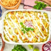 A baked casserole topped with melted cheese, white sauce, cilantro, and red onion slices, surrounded by bowls of rice, beans, lime wedges, cheese, and chopped vegetables on a festive tabletop.