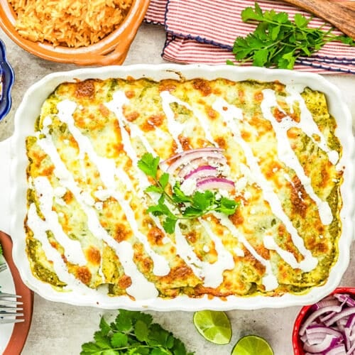 A baked casserole topped with melted cheese, white sauce, cilantro, and red onion slices, surrounded by bowls of rice, beans, lime wedges, cheese, and chopped vegetables on a festive tabletop.