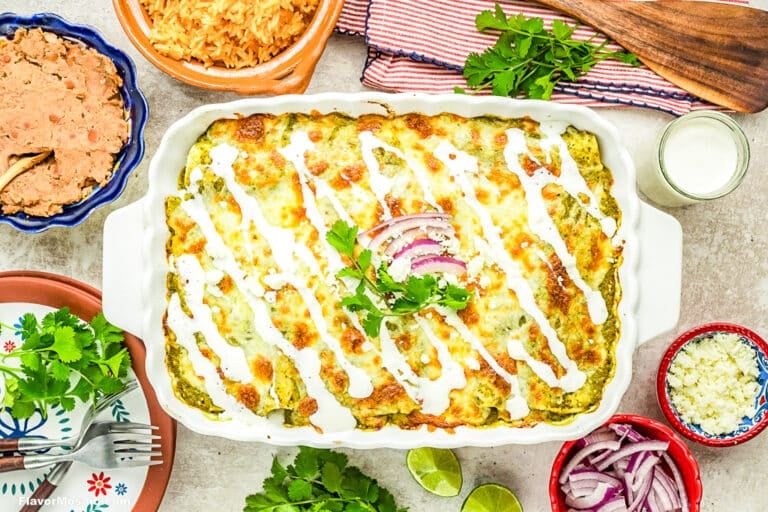A baked casserole topped with melted cheese, white sauce, cilantro, and red onion slices, surrounded by bowls of rice, beans, lime wedges, cheese, and chopped vegetables on a festive tabletop.