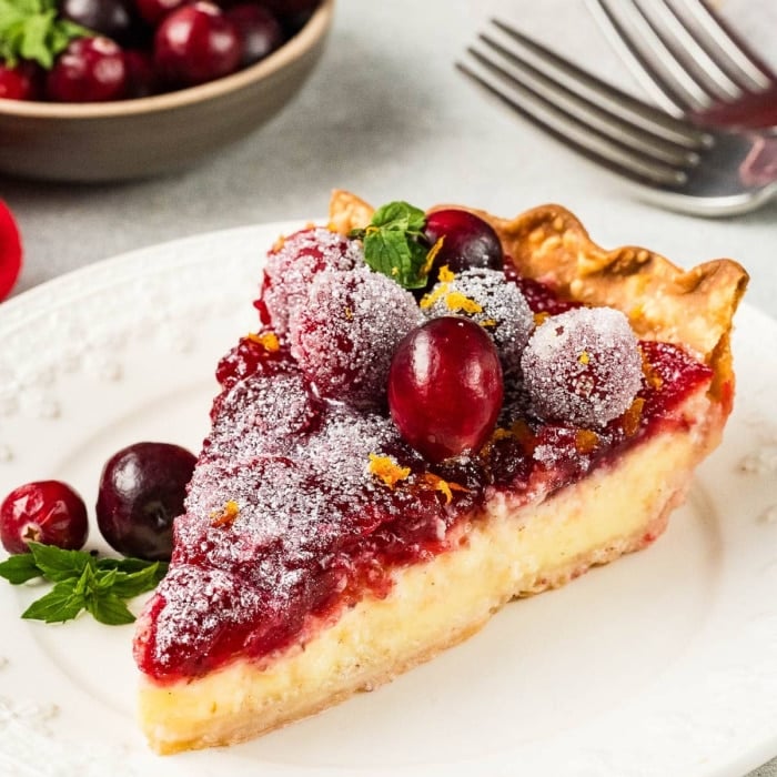 A slice of cranberry pie topped with sugared cranberries and orange zest sits on a white plate, garnished with mint. A bowl of fresh cranberries and forks are in the background.