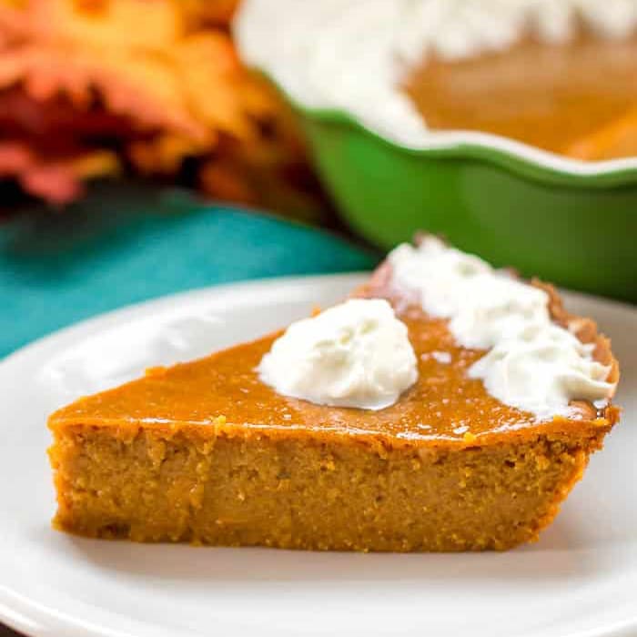 A slice of pumpkin pie topped with whipped cream sits on a white plate, with a pie dish and autumn leaves in the blurred background.