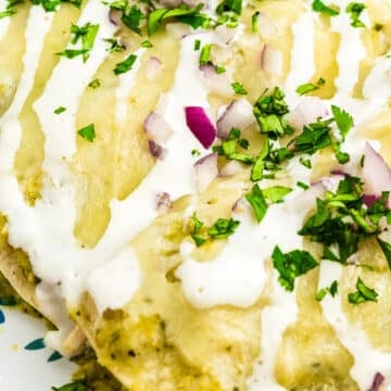 Close-up of enchiladas topped with melted cheese, drizzled with white sauce, and garnished with chopped red onion and fresh cilantro on a floral-patterned plate.