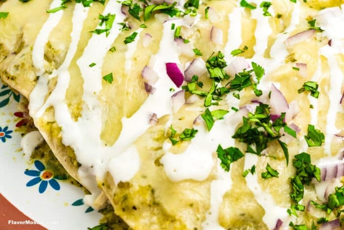 Close-up of enchiladas topped with melted cheese, drizzled with white sauce, and garnished with chopped red onion and fresh cilantro on a floral-patterned plate.