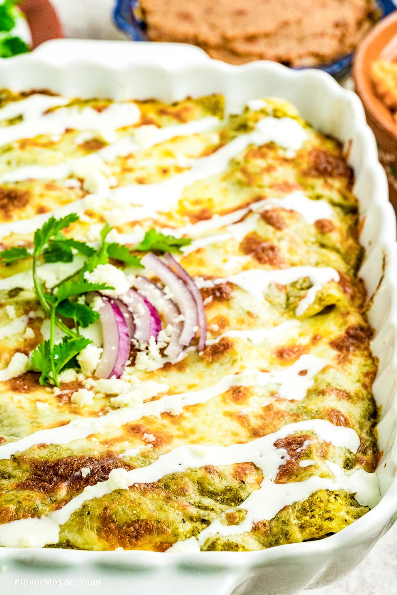 A close-up of a baked dish with green sauce, melted cheese, and drizzled white crema, garnished with cilantro and red onion slices in a white casserole dish. Tortillas and another blue dish are visible in the background.