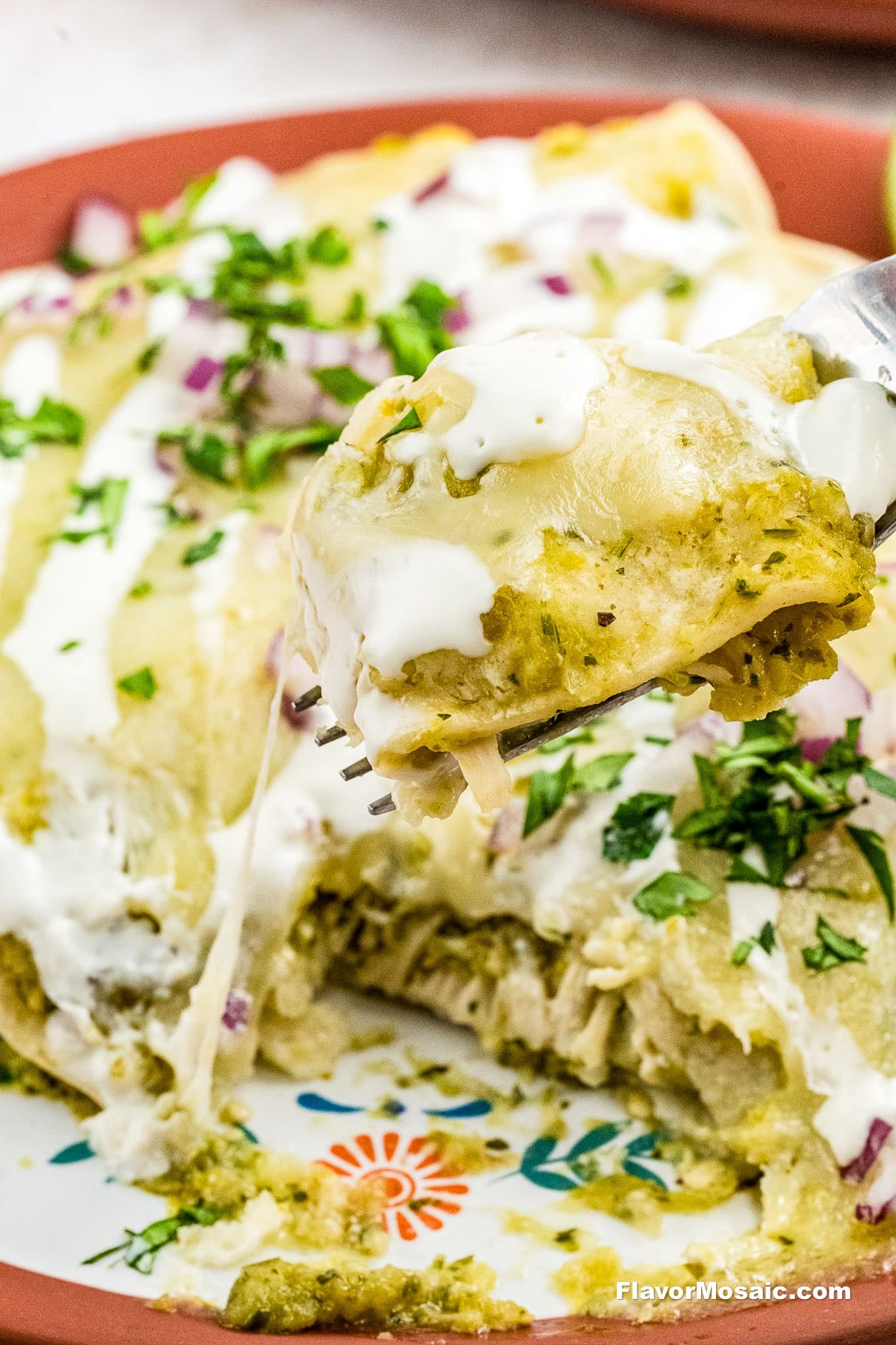 A close-up of a fork holding a cheesy chicken enchilada from a plate of enchiladas verdes, topped with green sauce, sour cream, chopped cilantro, and red onion. More enchiladas are visible in the background.