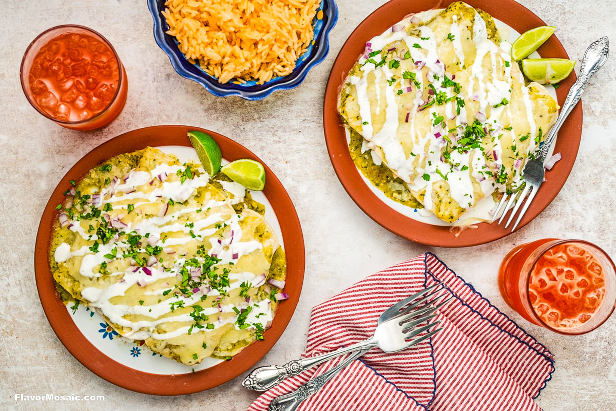 Two plates of enchiladas topped with sauce, chopped herbs, red onions, and drizzle of crema, garnished with lime wedges. Served with a side of Mexican rice and two glasses of a red drink, with utensils and a red striped napkin.
