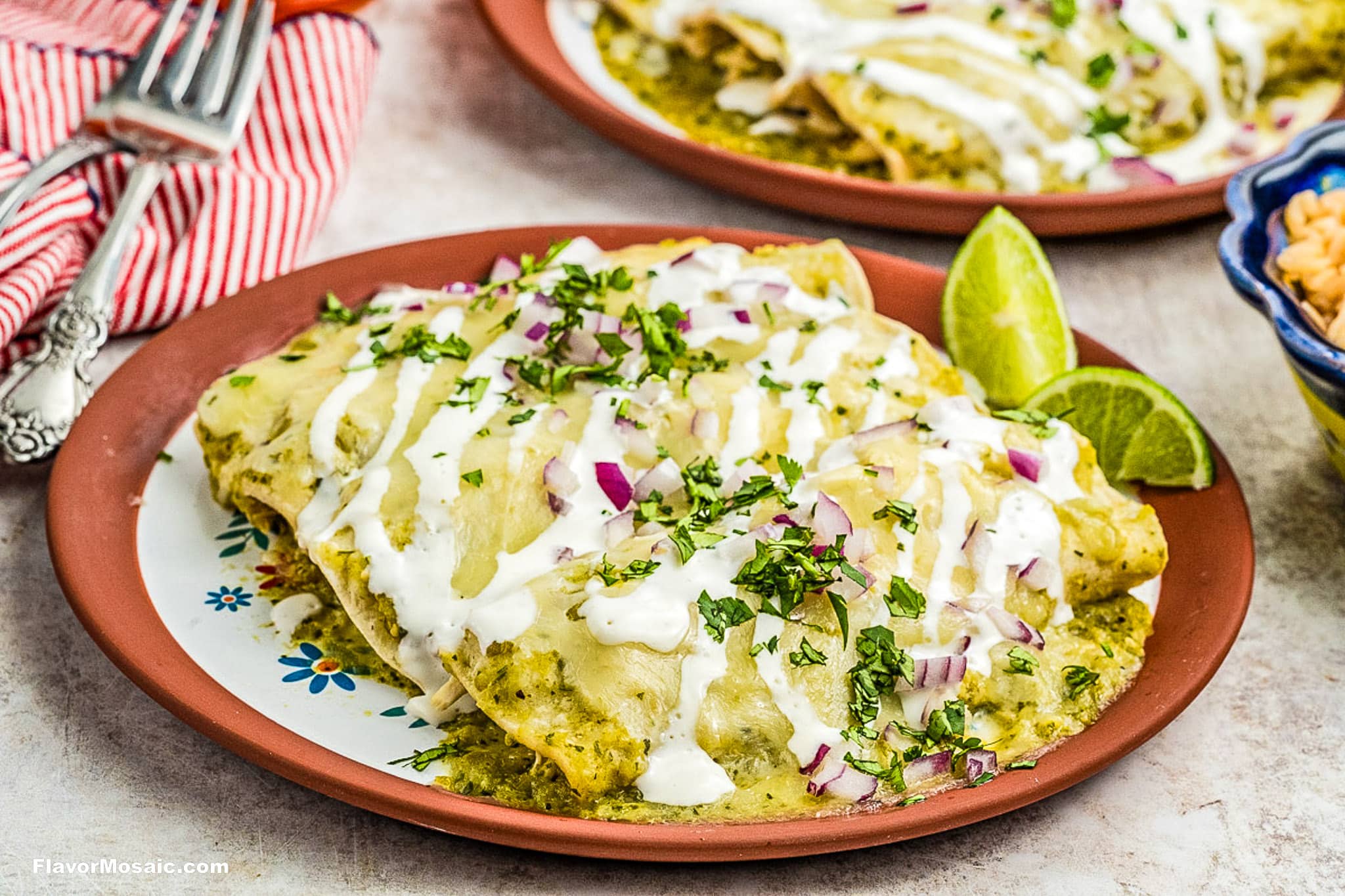 A plate of chicken enchiladas covered in green salsa, drizzled with sour cream, and topped with chopped cilantro and red onions, with lime wedges on the side. Another plate of enchiladas is in the background.
