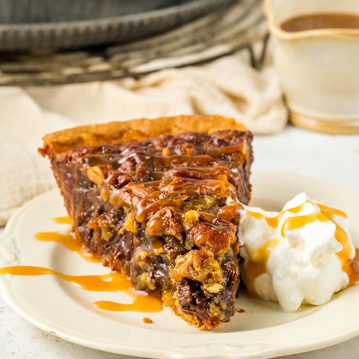 A slice of pecan pie topped with caramel drizzle sits on a plate next to a dollop of whipped cream, also drizzled with caramel, with a cup of coffee in the background.