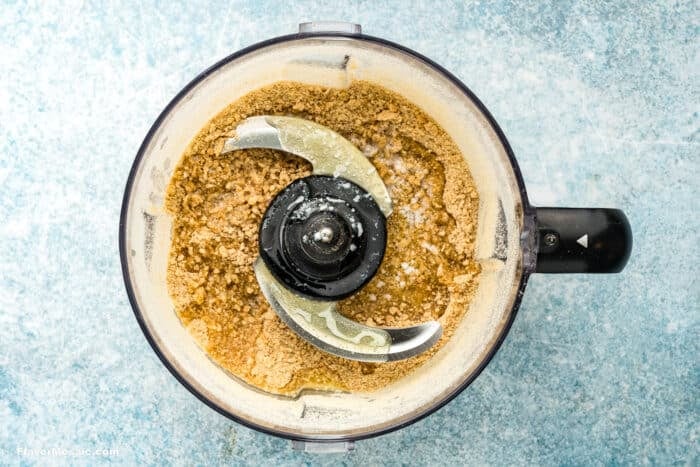 A food processor with partially blended nuts, turning into nut butter for a Key Lime Pie, sits on a light blue textured surface. The mixture appears coarse and slightly creamy, surrounding the metal blade inside the clear container.