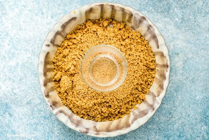 A pie pan filled with a layer of crumbly graham cracker crust, perfect for Key Lime Pie, with a small glass cup pressing down in the center to flatten the crumbs against the bottom of the pan.