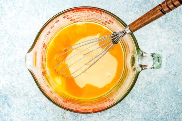 A glass measuring cup filled with beaten eggs and a metal whisk with a wooden handle, ready for making Key Lime Pie, placed on a light blue textured surface.