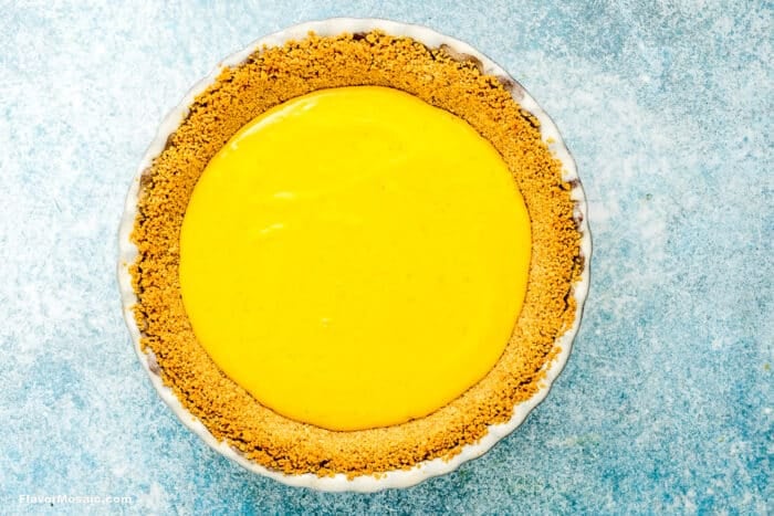A Key Lime Pie with a golden graham cracker crust and smooth, bright yellow filling, viewed from above on a light blue textured surface.