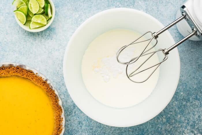 A bowl of cream and powdered sugar with a hand mixer sits beside a classic Key Lime Pie with a graham cracker crust and a small bowl of sliced limes, all arranged on a blue surface.