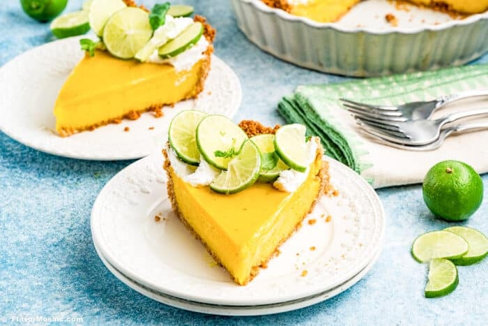 A slice of yellow Key Lime Pie with a crumb crust, topped with whipped cream and fresh lime slices, is served on a white plate. More Key Lime Pie, limes, and a fork are visible in the background.