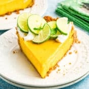 A slice of Key Lime Pie topped with whipped cream and fresh lime slices sits on a white plate, with a green napkin and whole limes in the background.