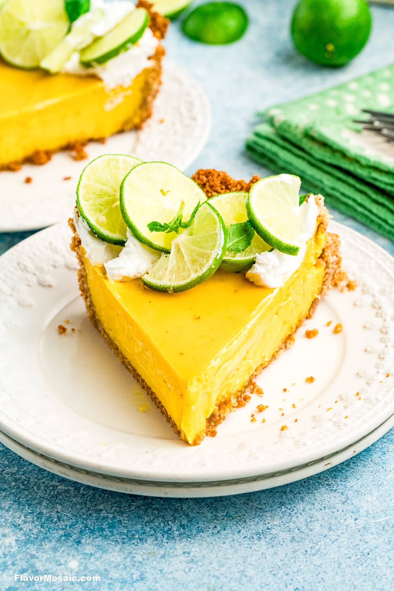 A slice of Key Lime Pie topped with whipped cream and fresh lime slices sits on a white plate, with a green napkin and whole limes in the background.