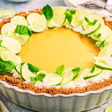 A classic Key Lime Pie in a fluted dish, topped with whipped cream, fresh lime slices, and mint leaves. The pie’s graham cracker crust sits on a light surface with plates and forks in the background.