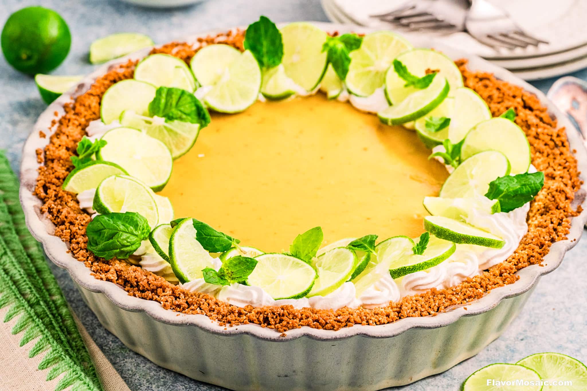 A classic Key Lime Pie in a fluted dish, topped with whipped cream, fresh lime slices, and mint leaves. The pie's graham cracker crust sits on a light surface with plates and forks in the background.