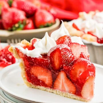 A slice of strawberry pie topped with whipped cream sits on a white plate. The pie is filled with fresh strawberries and a glossy red glaze, with more whole strawberries and pie in the background.
