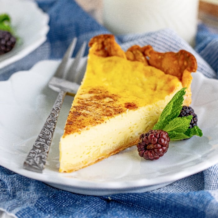 A slice of custard pie on a white plate, garnished with two blackberries and a sprig of mint. A fork rests beside the pie, and part of a blue cloth is visible underneath the plate.