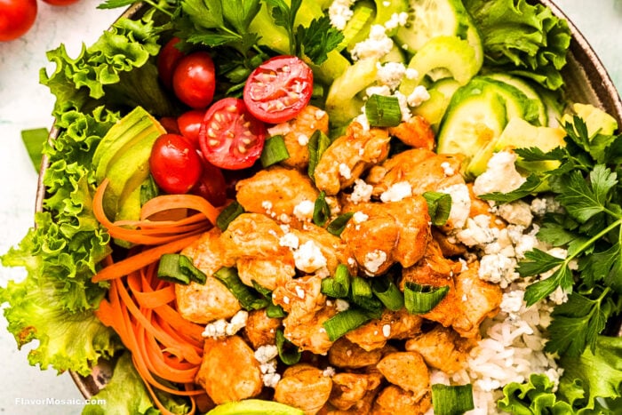 A colorful bowl with lettuce, sliced cucumbers, cherry tomatoes, carrots, buffalo chicken, white rice, blue cheese crumbles, and parsley, arranged in a vibrant, fresh display.