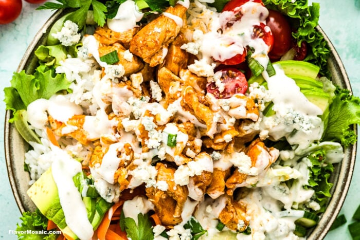 A close-up of a bowl filled with buffalo chicken, white rice, cherry tomatoes, avocado slices, lettuce, blue cheese crumbles, and creamy ranch dressing, garnished with fresh parsley.