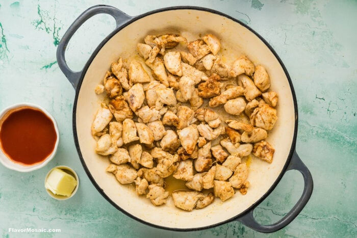 Chunks of cooked, seasoned chicken in a large round pot, with a small bowl of hot sauce and a container of butter next to it, on a light green textured surface.