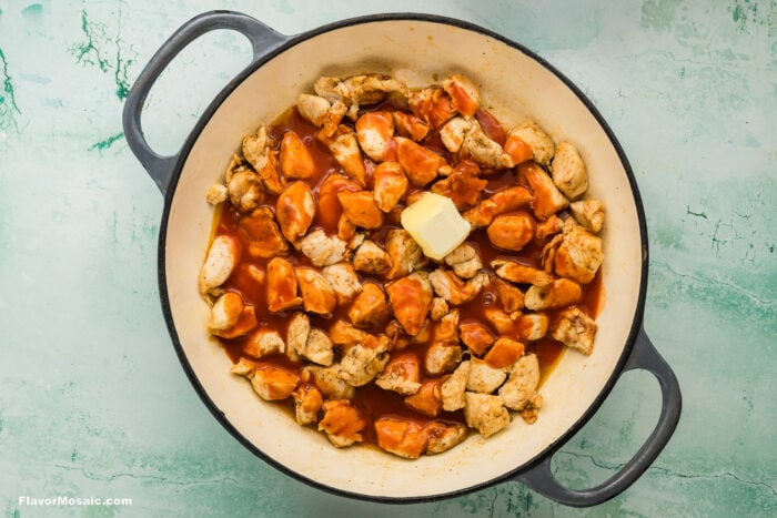 A round pot filled with cooked chicken pieces covered in orange sauce, topped with a pat of butter, on a light green textured background.