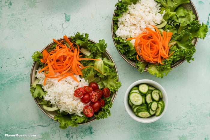 Two bowls with white rice, lettuce, grated carrot, cucumber slices, and cherry tomatoes on a green textured background, next to a small bowl of sliced cucumber.