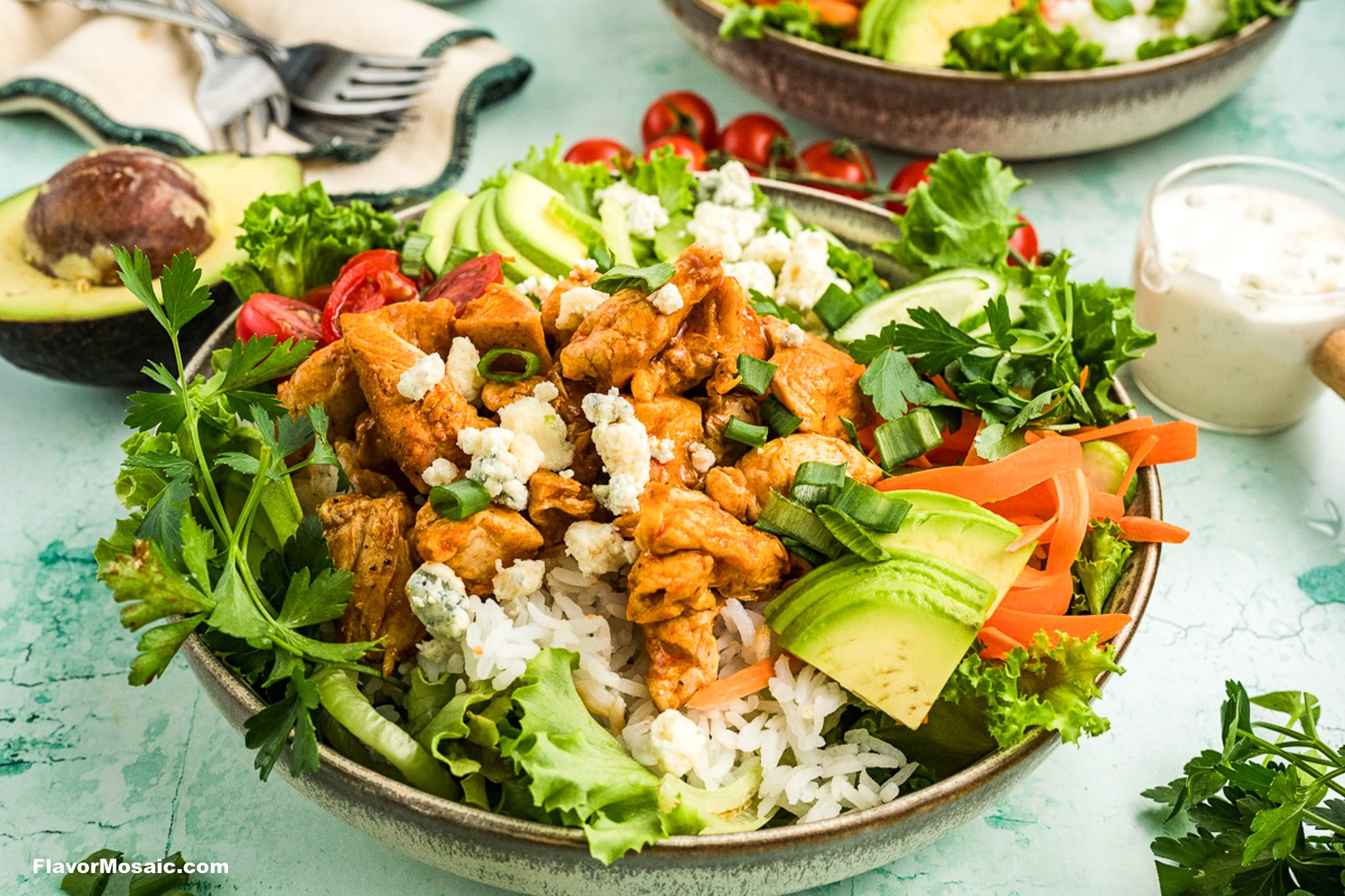 A bowl filled with rice, grilled chicken, avocado slices, lettuce, tomatoes, blue cheese crumbles, and fresh herbs, with a fork, avocado, and more salad bowls in the background on a green surface.