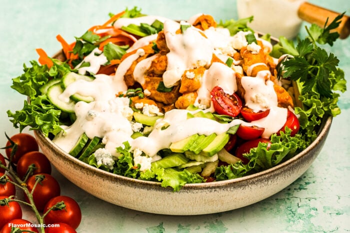 A bowl of fresh salad with leafy greens, cherry tomatoes, cucumber slices, carrot ribbons, crumbled blue cheese, and crispy fried chicken pieces, topped with creamy ranch dressing. Tomatoes and a scoop are on the side.