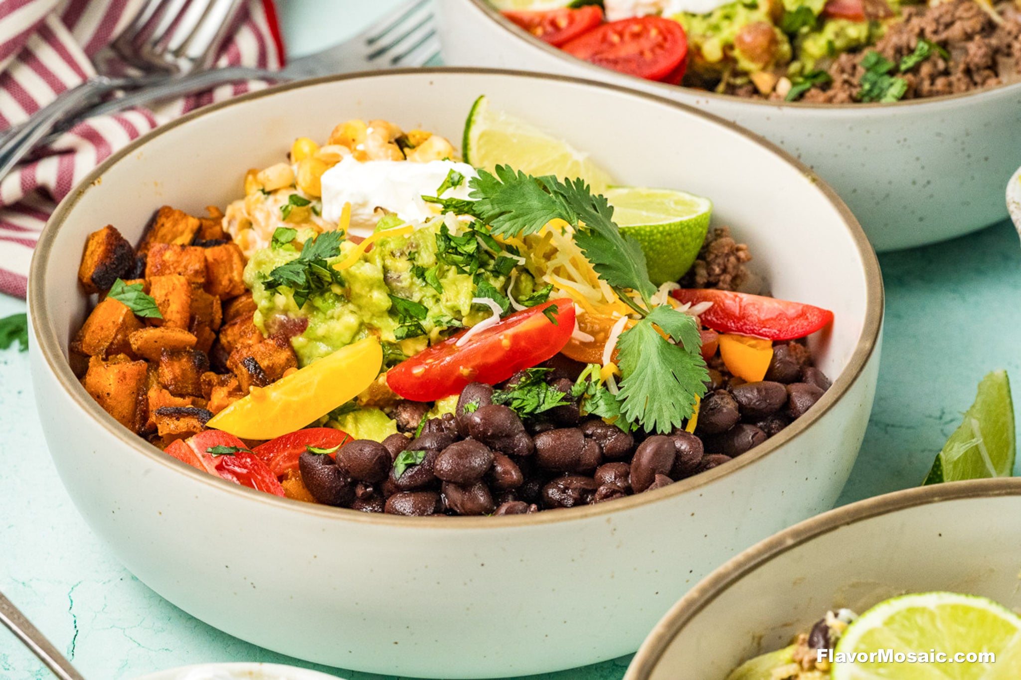 A vibrant Ground Beef Sweet Potato Bowl featuring black beans, roasted sweet potatoes, corn, guacamole, shredded cheese, cherry tomatoes, lime wedges, sour cream, bell peppers, and fresh cilantro artfully arranged in colorful sections.