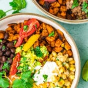 Two bowls filled with colorful burrito ingredients—black beans, ground beef sweet potato bowl mix, corn, guacamole, sour cream, shredded cheese, tomatoes, cilantro, and lime wedges—are arranged on a light blue background.