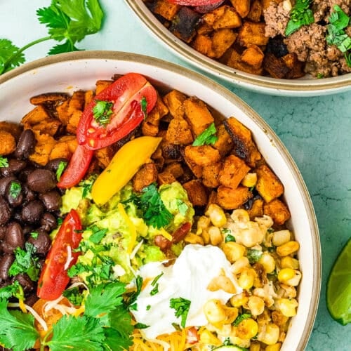 Two bowls filled with colorful burrito ingredients-black beans, ground beef sweet potato bowl mix, corn, guacamole, sour cream, shredded cheese, tomatoes, cilantro, and lime wedges-are arranged on a light blue background.