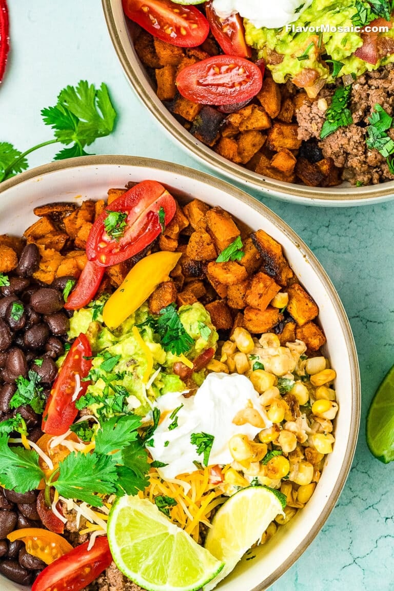 Two bowls filled with colorful burrito ingredients-black beans, ground beef sweet potato bowl mix, corn, guacamole, sour cream, shredded cheese, tomatoes, cilantro, and lime wedges-are arranged on a light blue background.