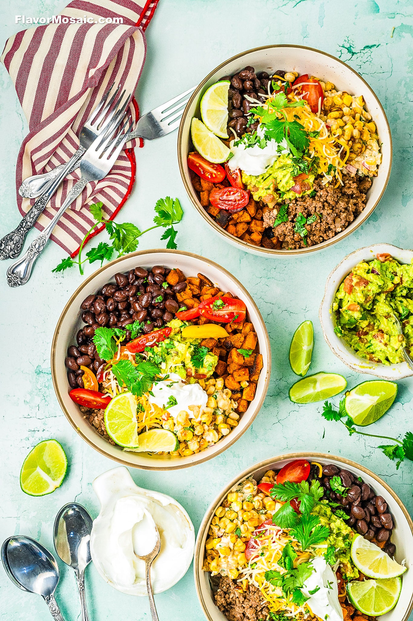 Three colorful Ground Beef Sweet Potato Bowls filled with ground meat, black beans, corn, cherry tomatoes, guacamole, sour cream, shredded cheese, and lime wedges, surrounded by extra toppings, utensils, and a striped napkin.