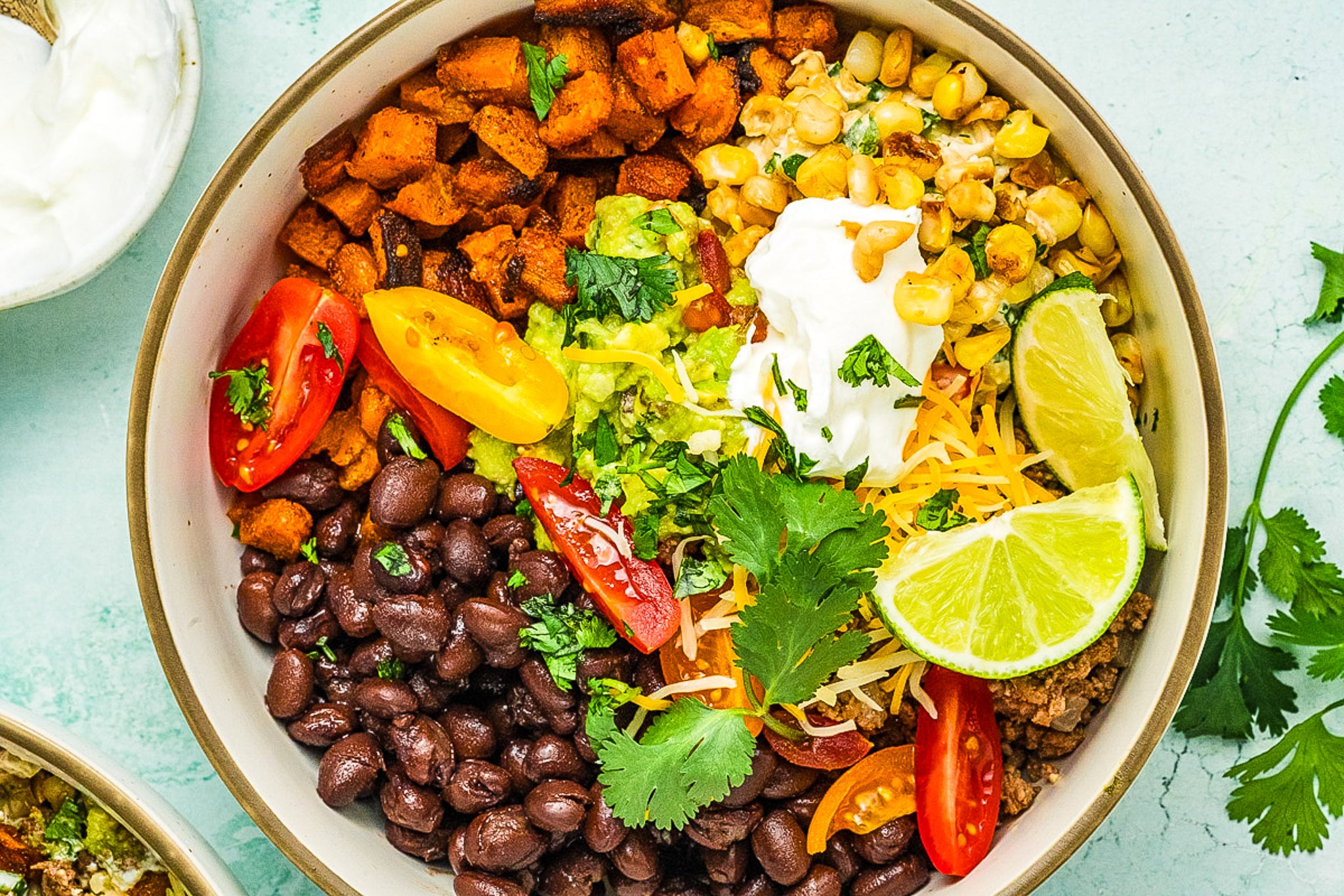 A colorful Ground Beef Sweet Potato Bowl filled with black beans, roasted sweet potatoes, corn, shredded cheese, cherry tomatoes, guacamole, sour cream, lime wedges, and cilantro on a light surface.