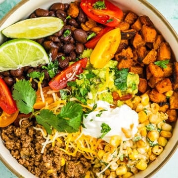 A vibrant Ground Beef Sweet Potato Bowl featuring black beans, roasted sweet potatoes, corn, shredded cheese, guacamole, sour cream, cherry tomatoes, lime wedges, and fresh cilantro in a round ceramic bowl.