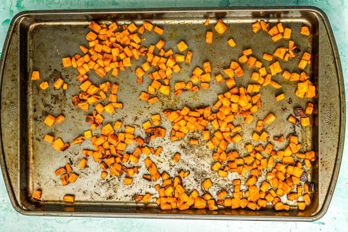 A baking sheet with scattered small roasted sweet potato cubes, lightly browned and caramelized, perfect for adding to a Ground Beef Sweet Potato Bowl. The tray is worn and slightly stained, set against a light turquoise background.