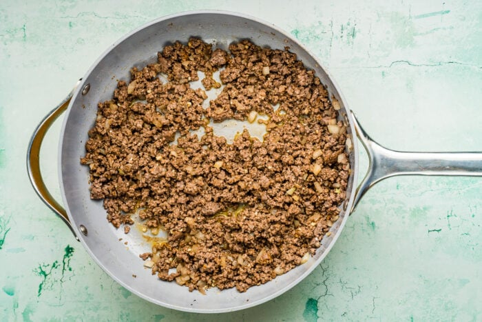 Ground beef cooked with small pieces of onion in a large silver skillet, set on a light green textured background-perfect as the base for a hearty Ground Beef Sweet Potato Bowl.