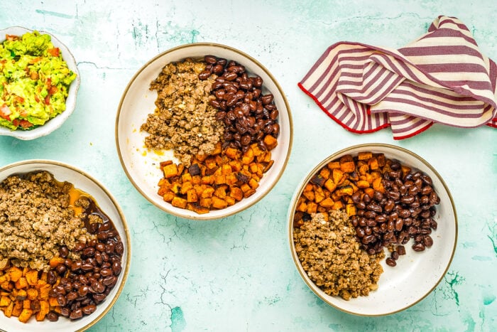 Three Ground Beef Sweet Potato Bowls sit on a light blue surface, each filled with black beans, seasoned ground meat, and roasted sweet potatoes. A bowl of guacamole and a red-and-white striped towel are also visible.