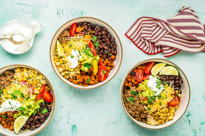 Three bowls of taco salad with ground beef, black beans, corn, tomatoes, shredded cheese, guacamole, and greens on a light blue surface evoke the flavors of a Ground Beef Sweet Potato Bowl. Served with lime wedges and sour cream for extra zest.