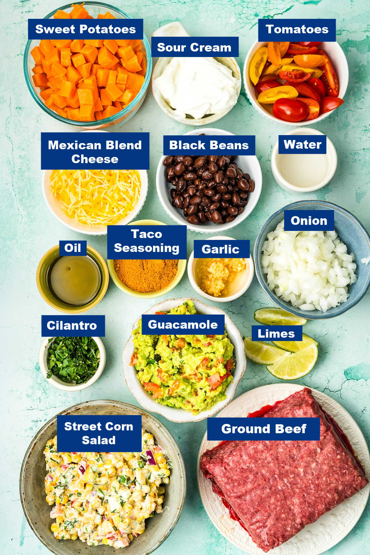 Ingredients for a Ground Beef Sweet Potato Bowl arranged on a light blue background, including sweet potatoes, sour cream, tomatoes, cheese, black beans, water, oil, taco seasoning, garlic, onion, cilantro, guacamole, limes and corn salad.