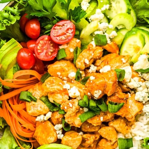 A colorful bowl filled with lettuce, cherry tomatoes, cucumber, sliced avocado, shredded carrots, rice, and seasoned chicken topped with blue cheese and chopped green onions. Fresh parsley garnishes the bowl.